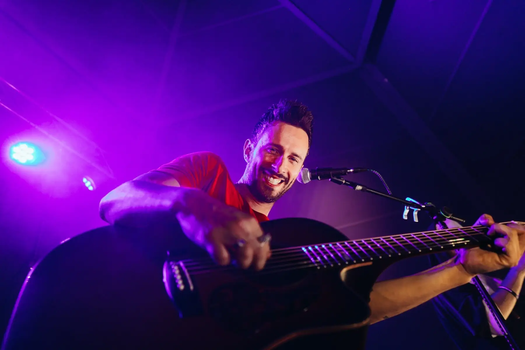 Musicista sorridente che suona la chitarra acustica sul palco durante la riapertura di un concerto.