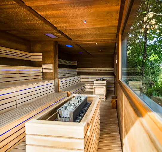 Sauna in legno con stufa di pietre e ampie vetrate. Ambiente rilassante e luminoso.